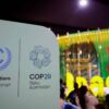 Cartel de la COP29 durante la Conferencia de la ONU sobre el Cambio Climático: a la izquierda, el emblema de la UNFCCC, con una letra "C" azul rodeada de ramas de olivo; a la derecha, el logotipo de la COP29 con hojas dispuestas en espiral. Debajo se lee "COP29 Baku Azerbaijan". Al fondo se distingue el pabellón de Brasil, con colores vibrantes, frente al cual se observan varias personas desenfocadas. COP30
