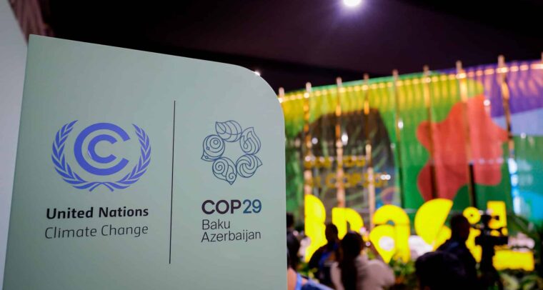 Cartel de la COP29 durante la Conferencia de la ONU sobre el Cambio Climático: a la izquierda, el emblema de la UNFCCC, con una letra "C" azul rodeada de ramas de olivo; a la derecha, el logotipo de la COP29 con hojas dispuestas en espiral. Debajo se lee "COP29 Baku Azerbaijan". Al fondo se distingue el pabellón de Brasil, con colores vibrantes, frente al cual se observan varias personas desenfocadas. COP30