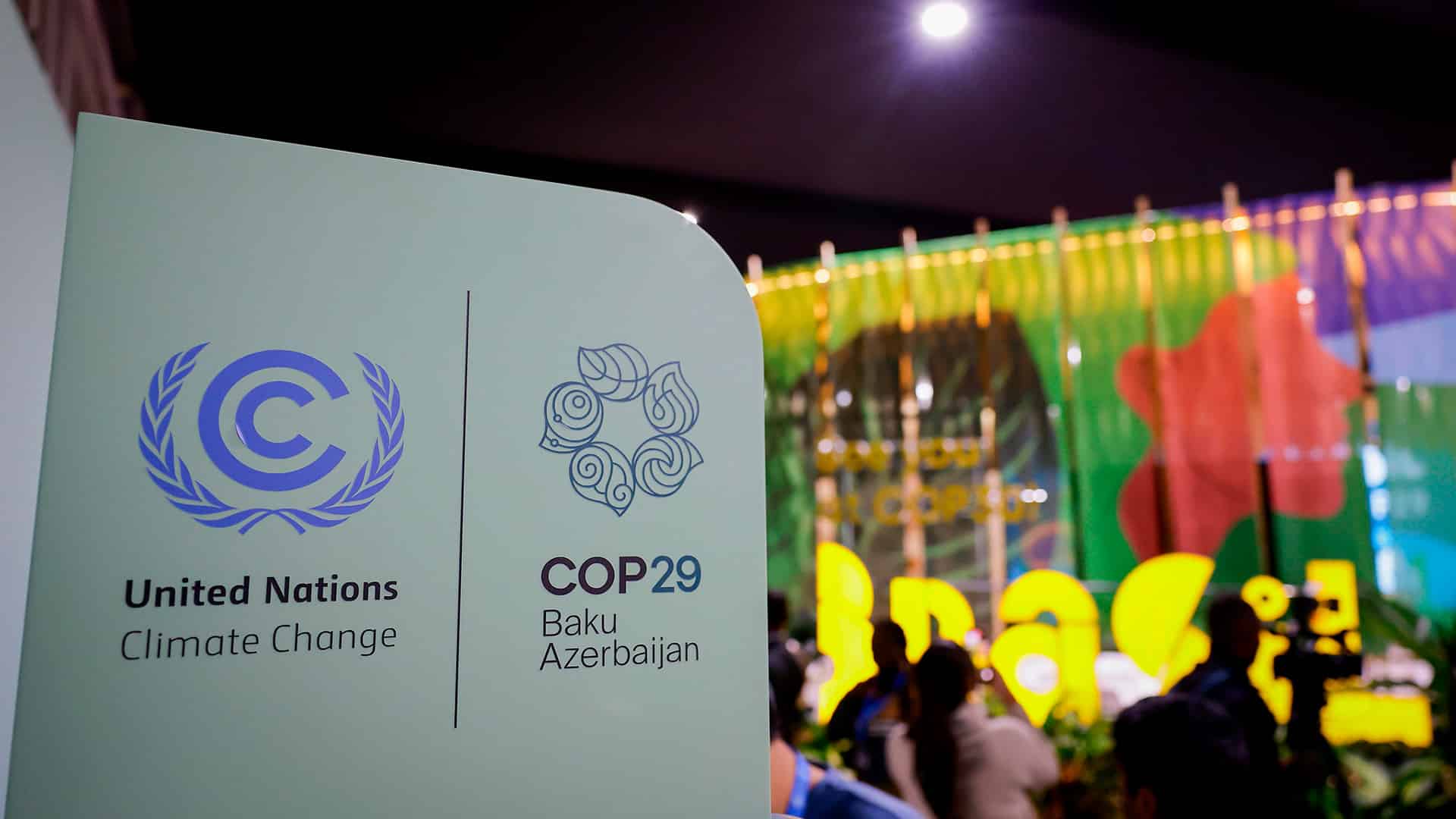 Cartel de la COP29 durante la Conferencia de la ONU sobre el Cambio Climático: a la izquierda, el emblema de la UNFCCC, con una letra "C" azul rodeada de ramas de olivo; a la derecha, el logotipo de la COP29 con hojas dispuestas en espiral. Debajo se lee "COP29 Baku Azerbaijan". Al fondo se distingue el pabellón de Brasil, con colores vibrantes, frente al cual se observan varias personas desenfocadas. COP30