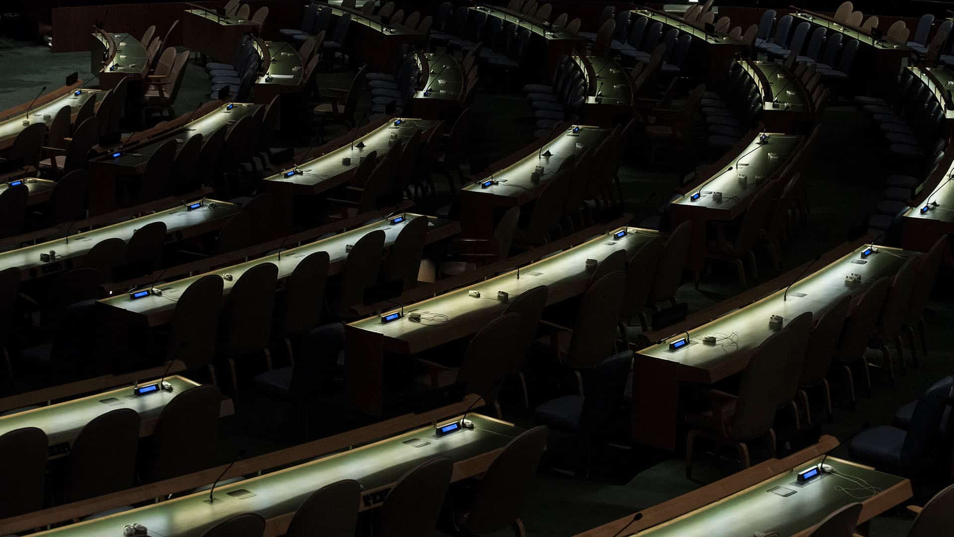 Salón de la Asamblea General de Naciones Unidas después de una reunión, sin delegados presentes, con la única iluminación procedente de los escritorios dispuestos en forma circular. ONU