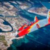 Dos aviones de la Patrulla Águila, con colores rojo, gris y amarillo, vuelan en formación sobre una zona costera con aguas turquesas y una urbanización, durante una exhibición aérea en el marco del Festival Aire 25 en la Academia General del Aire y del Espacio, el 4 de abril de 2025.