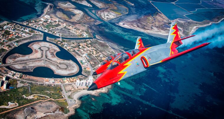 Two Patrulla Águila aircraft, painted in red, grey, and yellow, fly in formation over a coastal area with turquoise waters and a residential development, during an air show held as part of the Aire 25 Festival at the General Air and Space Academy on April 4, 2025.