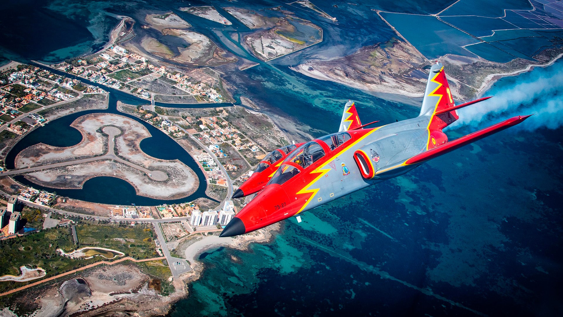 Dos aviones de la Patrulla Águila, con colores rojo, gris y amarillo, vuelan en formación sobre una zona costera con aguas turquesas y una urbanización, durante una exhibición aérea en el marco del Festival Aire 25 en la Academia General del Aire y del Espacio, el 4 de abril de 2025.