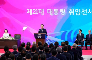 Ceremonia de investidura de Lee Jae-myung, como presidente número 21 de Corea del Sur en la Asamblea Nacional de Seúl, el 4 de junio de 2025. Lee está de pie en el podio presidencial, con traje oscuro y corbata azul, dirigiéndose a la audiencia. Al fondo, una pantalla rosa muestra el texto 'La ceremonia de investidura del 21.º presidente' en coreano, junto a banderas nacionales. Lee Jae-myung