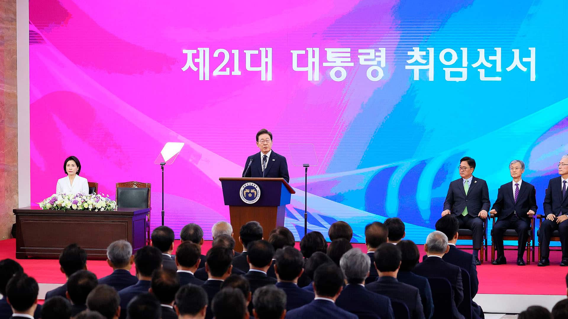 Ceremonia de investidura de Lee Jae-myung, como presidente número 21 de Corea del Sur en la Asamblea Nacional de Seúl, el 4 de junio de 2025. Lee está de pie en el podio presidencial, con traje oscuro y corbata azul, dirigiéndose a la audiencia. Al fondo, una pantalla rosa muestra el texto 'La ceremonia de investidura del 21.º presidente' en coreano, junto a banderas nacionales. Lee Jae-myung