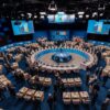 Vista panorámica del salón principal del World Forum de La Haya durante la Cena de Trabajo del Consejo OTAN-Ucrania en la Cumbre de la OTAN 2025. En el centro, destaca una gran mesa redonda con el emblema de la OTAN, rodeada por representantes. El espacio está flanqueado por pantallas informativas y una exposición de banderas nacionales. OTAN