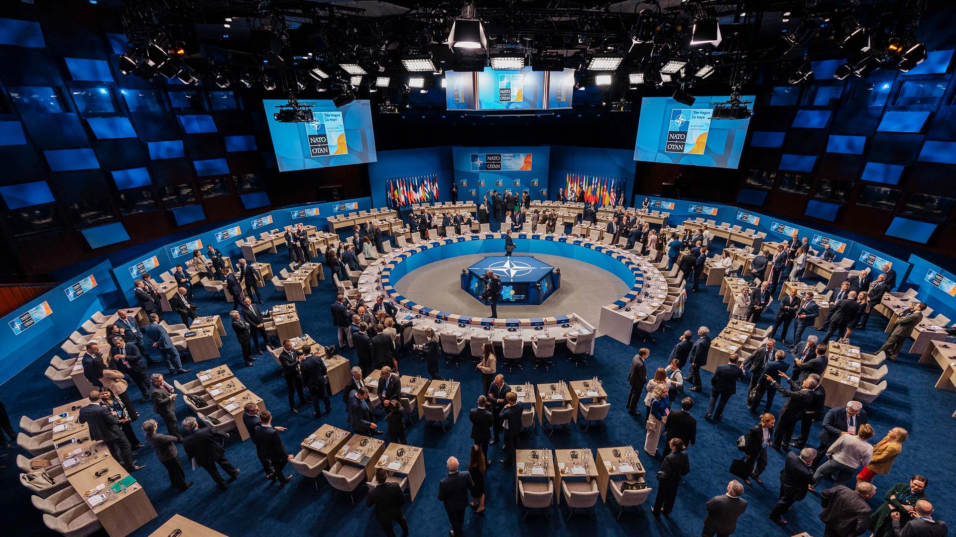 Vista panorámica del salón principal del World Forum de La Haya durante la Cena de Trabajo del Consejo OTAN-Ucrania en la Cumbre de la OTAN 2025. En el centro, destaca una gran mesa redonda con el emblema de la OTAN, rodeada por representantes. El espacio está flanqueado por pantallas informativas y una exposición de banderas nacionales. OTAN
