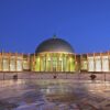 Exterior plaza of the Seville Conference and Exhibition Centre (FIBES) at dusk, featuring its illuminated dome, a symmetrical colonnade, and a glass façade. In the foreground, a mosaic with maps and the dates 1492–1992 commemorates the fifth centenary of the discovery of America. Aid