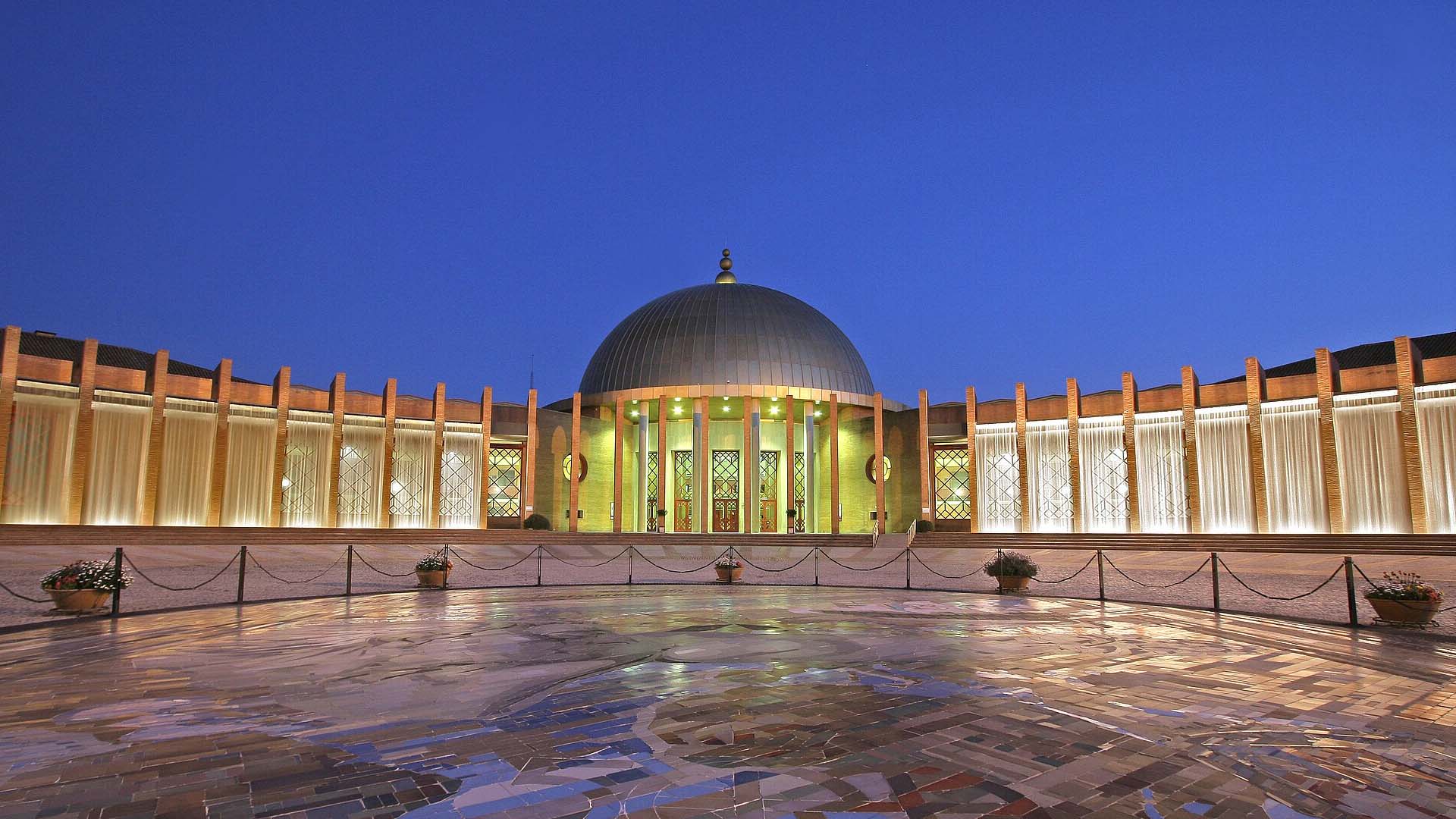 Exterior plaza of the Seville Conference and Exhibition Centre (FIBES) at dusk, featuring its illuminated dome, a symmetrical colonnade, and a glass façade. In the foreground, a mosaic with maps and the dates 1492–1992 commemorates the fifth centenary of the discovery of America. Aid
