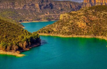 Panorámica del embalse de Camarasa (Cataluña), con aguas esmeralda rodeadas de montañas escarpadas y frondosos bosques, bajo un cielo azul que contrasta con algunas nubes. Agua