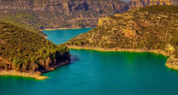Panorámica del embalse de Camarasa (Cataluña), con aguas esmeralda rodeadas de montañas escarpadas y frondosos bosques, bajo un cielo azul que contrasta con algunas nubes. Agua