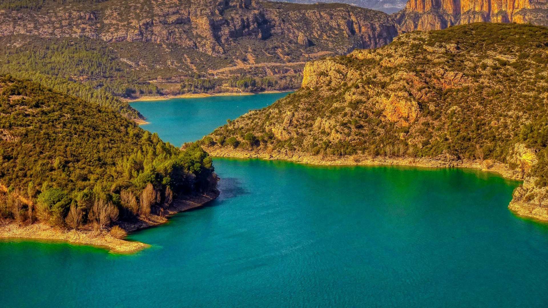 Panorámica del embalse de Camarasa (Cataluña), con aguas esmeralda rodeadas de montañas escarpadas y frondosos bosques, bajo un cielo azul que contrasta con algunas nubes. Agua