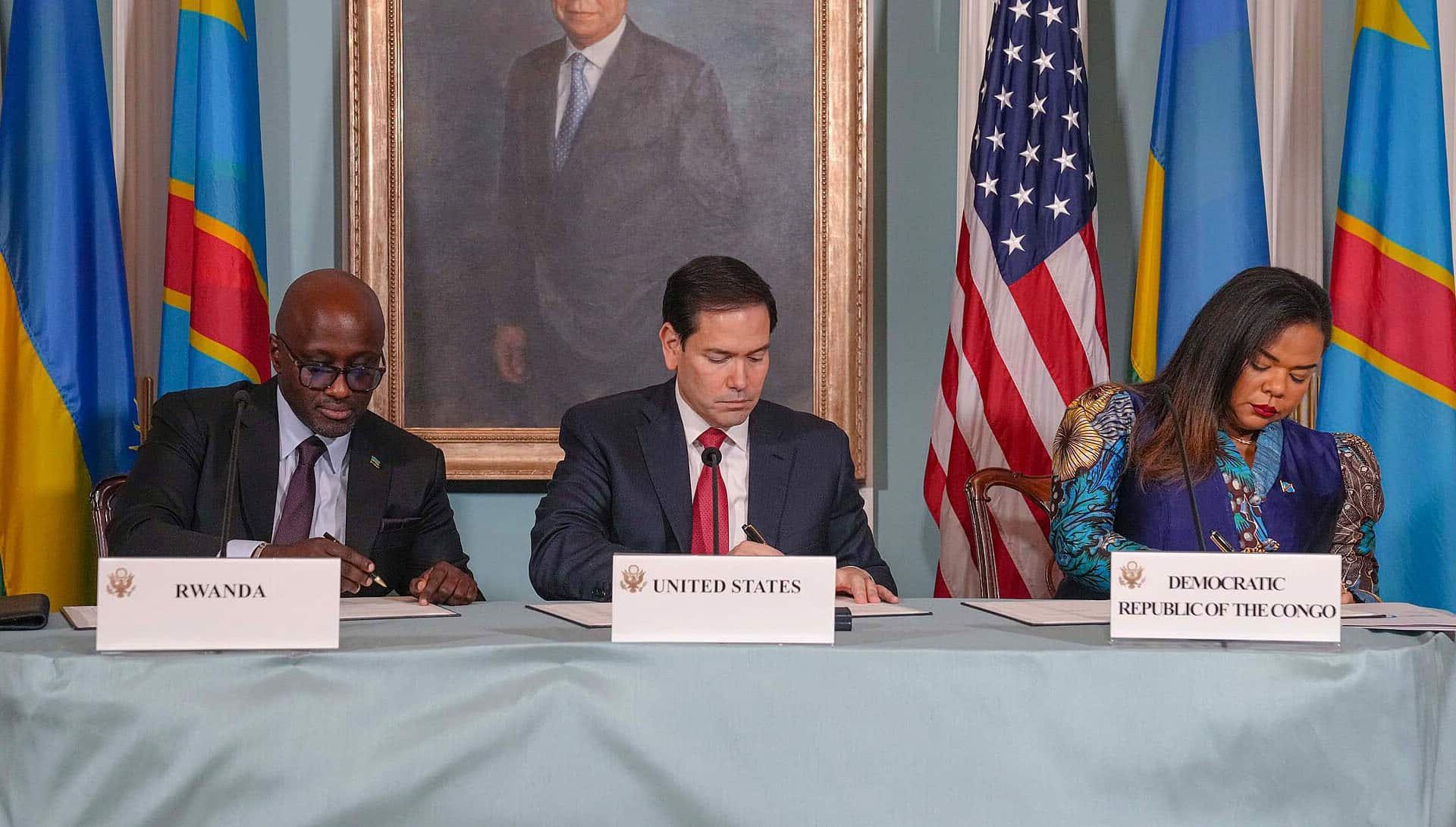Marco Rubio, la ministra de Asuntos Exteriores de la República Democrática del Congo, Thérèse Kayikwamba Wagner, y el ministro de Asuntos Exteriores de Ruanda, Olivier Nduhungirehe, sentados firmando el Acuerdo de Paz en el Departamento de Estado en Washington D.C., el 27 de junio de 2025. Frente a ellos, una mesa con los nombres de los tres países y banderas de la República Democrática del Congo, Ruanda y Estados Unidos a ambos lados. Acuerdo