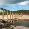 A long bridge with tall concrete pillars spans a nearly dry reservoir in Aceredo (Spain), revealing the sandy and rocky bed below. Sparse water remains in parts of the reservoir. In the background, green forested hills rise under a partly cloudy sky. Spain