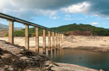 A long bridge with tall concrete pillars spans a nearly dry reservoir in Aceredo (Spain), revealing the sandy and rocky bed below. Sparse water remains in parts of the reservoir. In the background, green forested hills rise under a partly cloudy sky. Spain