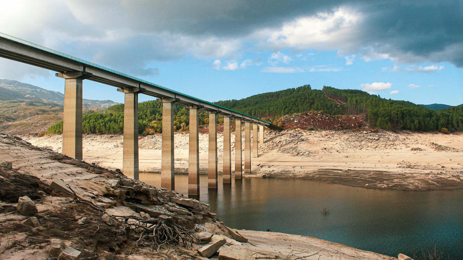 A long bridge with tall concrete pillars spans a nearly dry reservoir in Aceredo (Spain), revealing the sandy and rocky bed below. Sparse water remains in parts of the reservoir. In the background, green forested hills rise under a partly cloudy sky. Spain