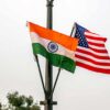 Las banderas nacionales de la India y de Estados Unidos ondean desde una farola de latón negro en Nueva Delhi bajo un día nublado. En la parte baja, se vislumbran copas de árboles verdes. India