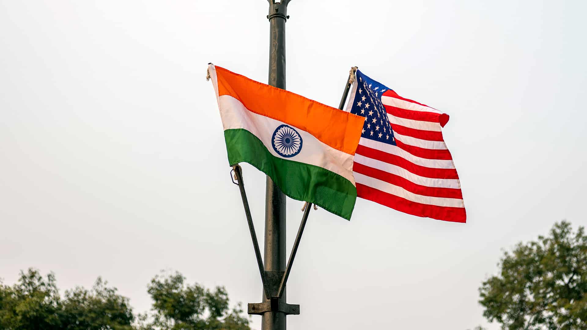 Las banderas nacionales de la India y de Estados Unidos ondean desde una farola de latón negro en Nueva Delhi bajo un día nublado. En la parte baja, se vislumbran copas de árboles verdes. India