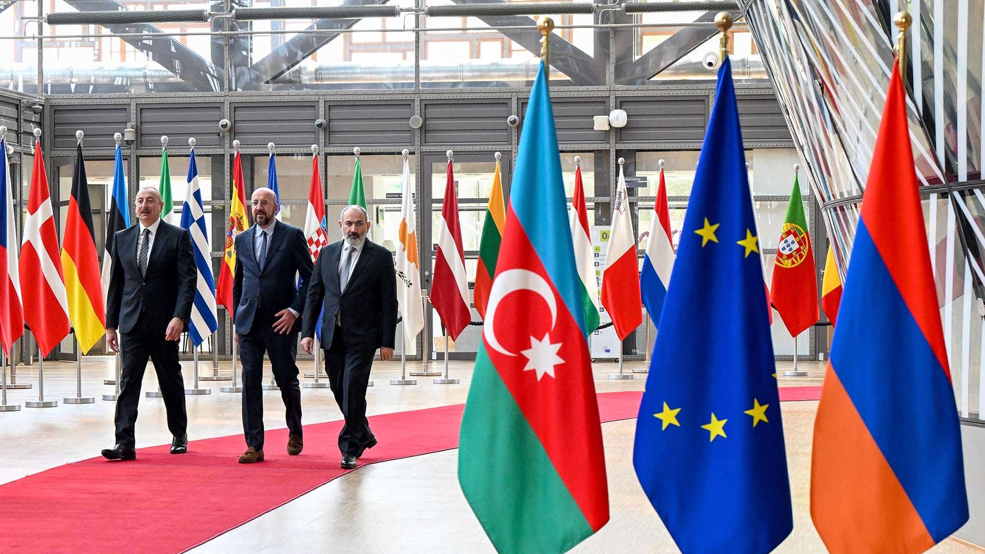 De izquierda a derecha: Ilham Aliyev (presidente de Azerbaiyán), Charles Michel (presidente del Consejo Europeo en 2023) y Nikol Pashinián (primer ministro de Armenia) caminan por la alfombra roja rodeada de banderas en la entrada de la sede del Consejo Europeo, para la reunión con los mandatarios de Francia y Alemania (1/06/2023), tras los enfrentamientos entre Azerbaiyán y Armenia en la región de Nagorno Karabaj. Se destacan las banderas de Azerbaiyán, la UE y Armenia en primer plano.