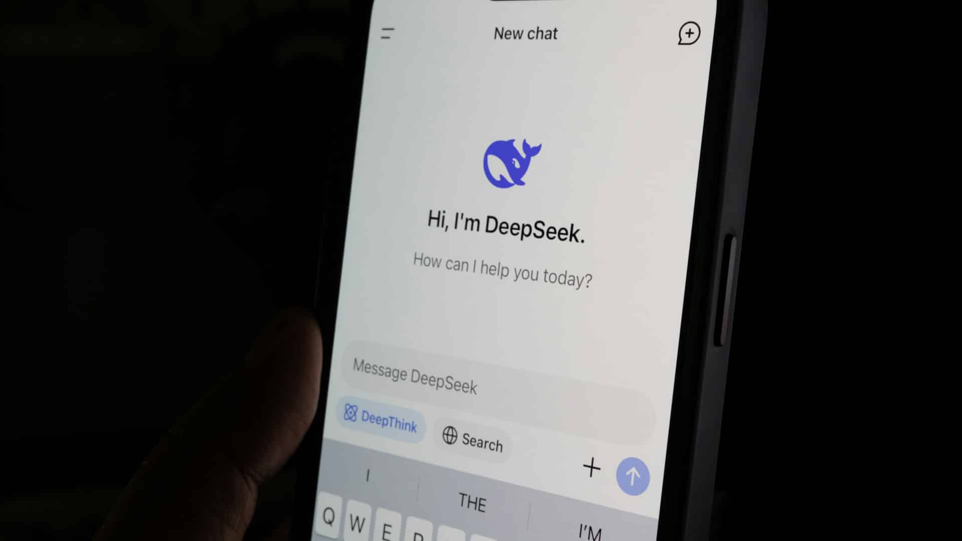 Una persona sostiene un móvil en su mano en un sitio oscuro. La pantalla iluminada muestra la ventana de bienvenida de la aplicación de DeepSeek en inglés con el mensaje “I'm DeepSeek. How can I help you today?” (Soy DeepSeek. ¿En qué puedo ayudarte hoy).