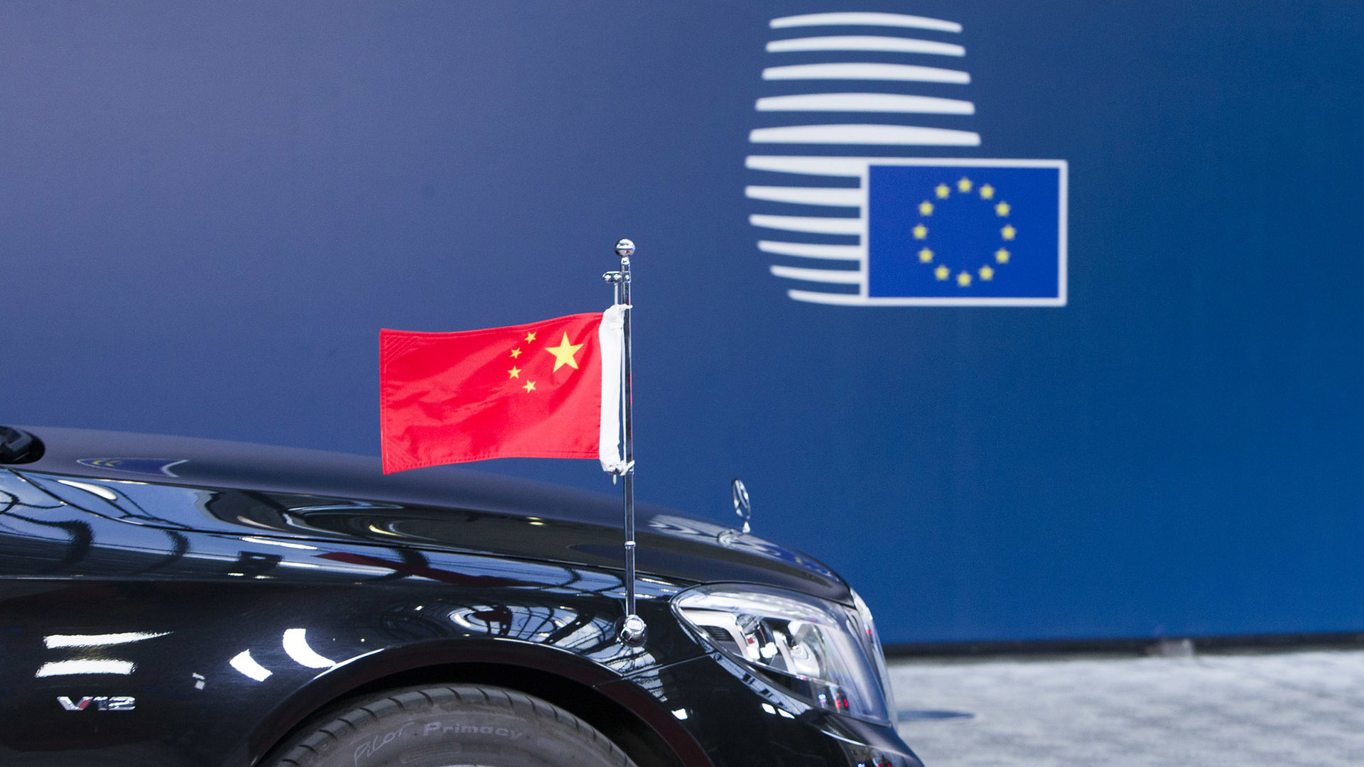 Parte frontal de un coche negro de lujo con una bandera de China en miniatura, frente a un fondo azul con el logo del Consejo Europeo. La foto es de la Cumbre China-UE de 2017 en Bruselas (Bélgica).