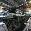 Ensamblaje de un carro de combate Lynx. dentro de una instalación industrial con techos altos, rodeado de escaleras metálicas, herramientas y un puente grúa amarillo marcado “Demag ZKKE”, en la empresa de defensa Rheinmetall en Alemania.