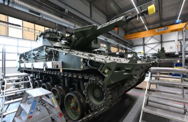 Ensamblaje de un carro de combate Lynx. dentro de una instalación industrial con techos altos, rodeado de escaleras metálicas, herramientas y un puente grúa amarillo marcado “Demag ZKKE”, en la empresa de defensa Rheinmetall en Alemania.