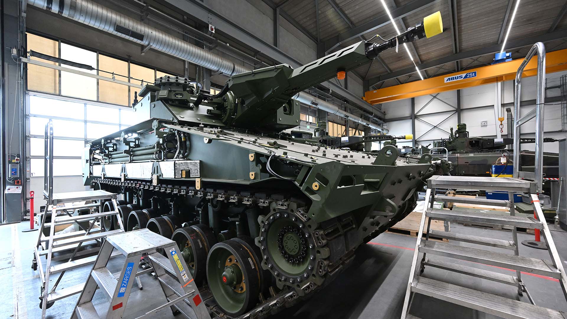 Ensamblaje de un carro de combate Lynx. dentro de una instalación industrial con techos altos, rodeado de escaleras metálicas, herramientas y un puente grúa amarillo marcado “Demag ZKKE”, en la empresa de defensa Rheinmetall en Alemania.
