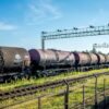 Vagones cisterna con petróleo ruso alineados sobre una vía férrea en una estación cerca de Ivanogrod (Rusia), con una cerca metálica en primer plano y una estructura metálica elevada al fondo. El cielo está parcialmente despejado y hay vegetación verde a los lados de las vías.