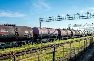 Vagones cisterna con petróleo ruso alineados sobre una vía férrea en una estación cerca de Ivanogrod (Rusia), con una cerca metálica en primer plano y una estructura metálica elevada al fondo. El cielo está parcialmente despejado y hay vegetación verde a los lados de las vías.