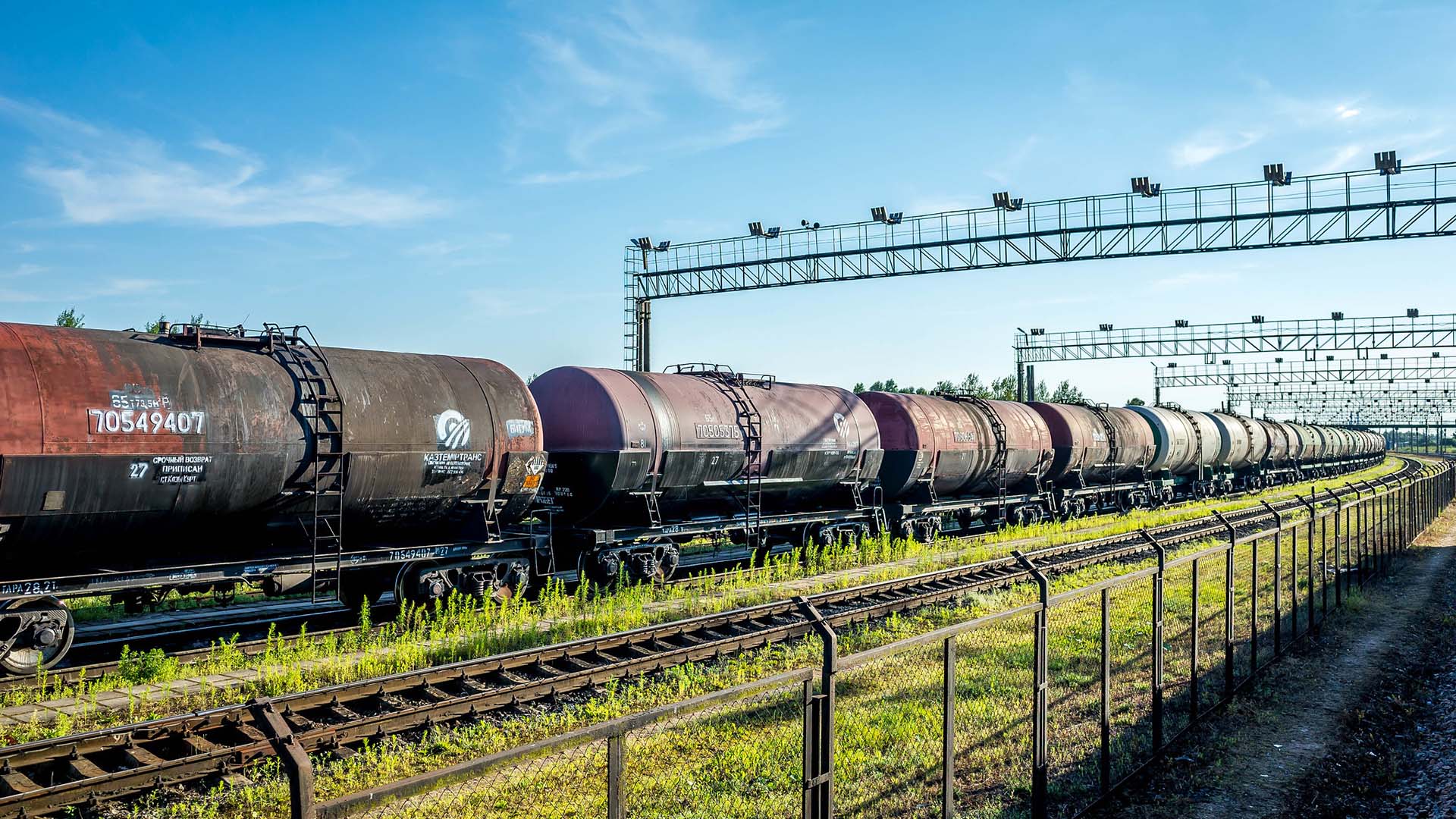 Vagones cisterna con petróleo ruso alineados sobre una vía férrea en una estación cerca de Ivanogrod (Rusia), con una cerca metálica en primer plano y una estructura metálica elevada al fondo. El cielo está parcialmente despejado y hay vegetación verde a los lados de las vías.