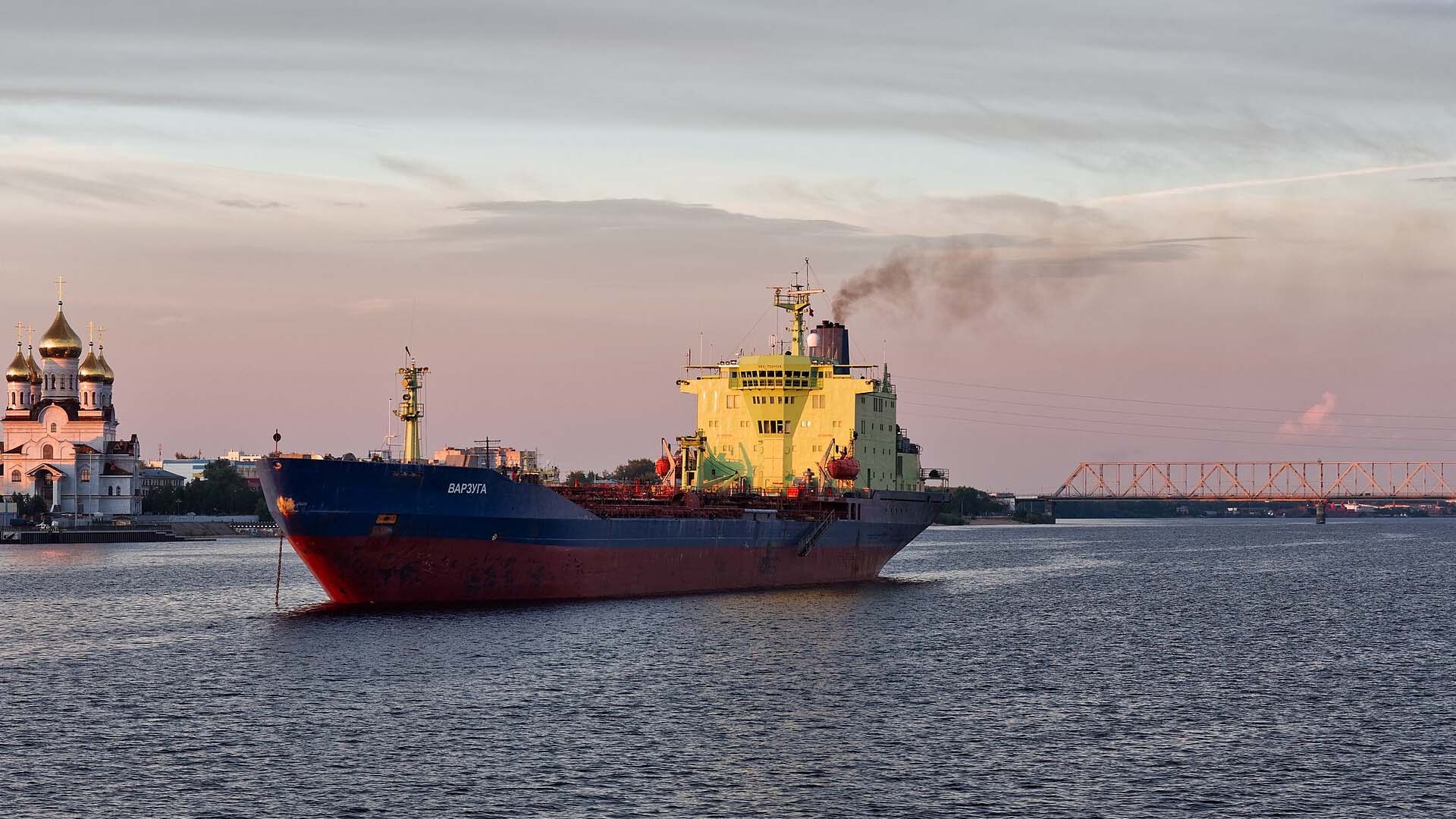 El petrolero Varzuga navega al atardecer por el río Dviná Septentrional en Arcángel (Rusia). A la izquierda se ve una iglesia ortodoxa con cúpulas doradas y al fondo un puente cruza el río. Flota fantasma
