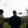 Three people with their backs turned, two in military uniform and one in a suit, watch two military helicopters flying over a green field with trees in the background. Prime Minister Keir Starmer visits a military training camp in South West England with New Zealand Prime Minister Christopher Luxon.