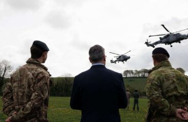 Three people with their backs turned, two in military uniform and one in a suit, watch two military helicopters flying over a green field with trees in the background. Prime Minister Keir Starmer visits a military training camp in South West England with New Zealand Prime Minister Christopher Luxon.