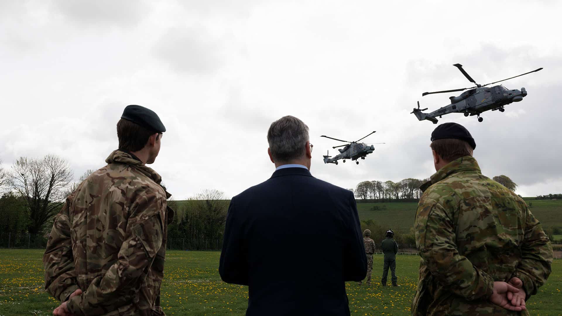 Three people with their backs turned, two in military uniform and one in a suit, watch two military helicopters flying over a green field with trees in the background. Prime Minister Keir Starmer visits a military training camp in South West England with New Zealand Prime Minister Christopher Luxon.