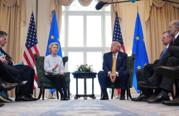 Ursula von der Leyen (presidenta de la Comisión Europea) y Donald Trump (presidente de EEUU) conversan frenta a frente con una mesa y arreglo floral entre ellos durante el encuentro bilateral en Turnberry (Escocia), el 27 de julio de 2025, acompañados por sus delegaciones a los lados y con banderas oficiales de la UE y EEUU al fondo.