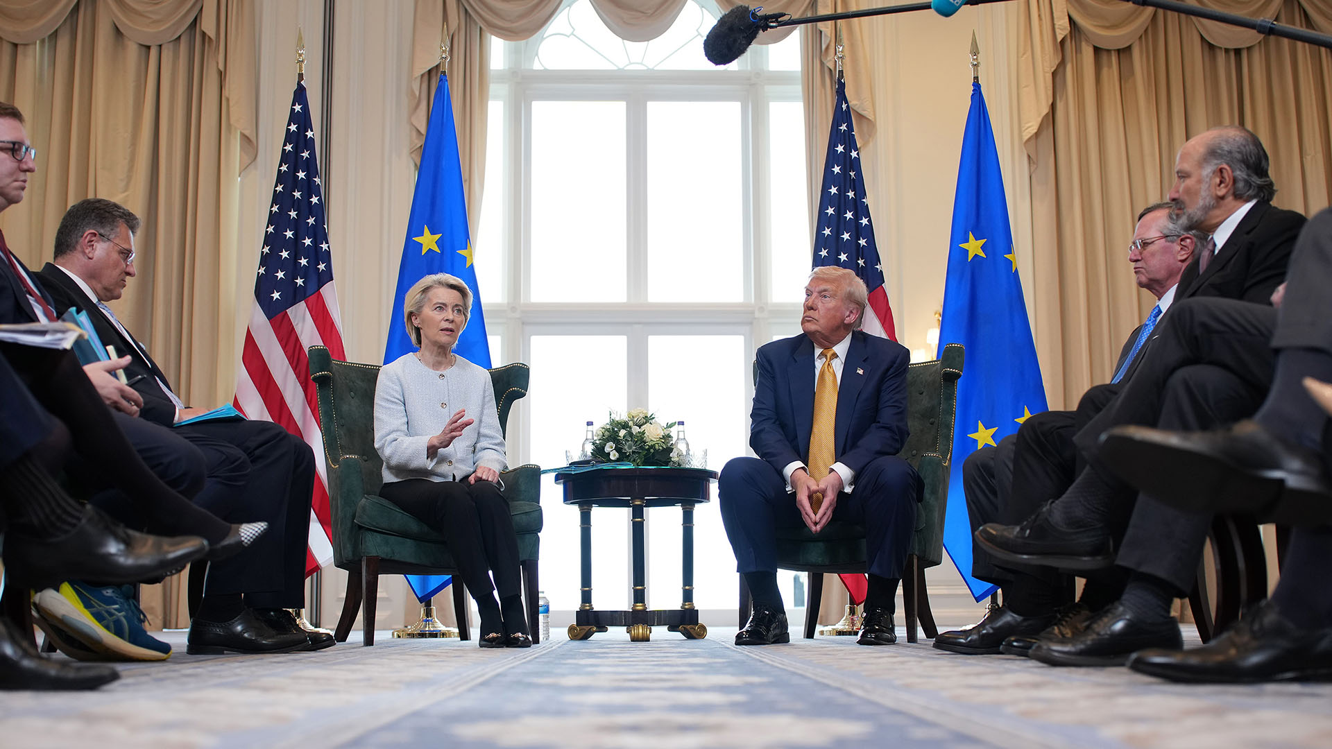 Ursula von der Leyen (presidenta de la Comisión Europea) y Donald Trump (presidente de EEUU) conversan frenta a frente con una mesa y arreglo floral entre ellos durante el encuentro bilateral en Turnberry (Escocia), el 27 de julio de 2025, acompañados por sus delegaciones a los lados y con banderas oficiales de la UE y EEUU al fondo.