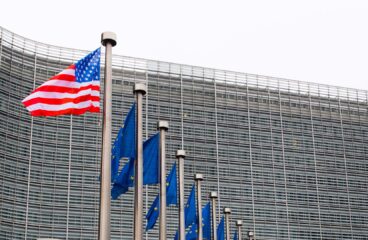 Fachada de la Comisión Europea en Bruselas con banderas de la Unión Europea alineadas y una bandera de Estados Unidos ondeando en primer plano en un día nublado. Acuerdo