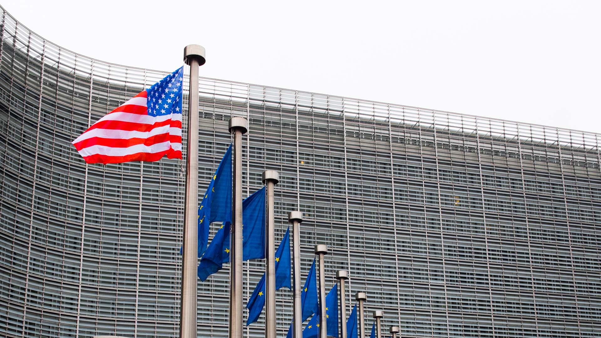 Fachada de la Comisión Europea en Bruselas con banderas de la Unión Europea alineadas y una bandera de Estados Unidos ondeando en primer plano en un día nublado. Acuerdo