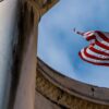 Perspectiva en contrapicado de la bandera de EEUU ondeando con firmeza entre columnas clásicas de piedra en Salt Lake City (Utah, EEUU), perfilada contra un cielo azul sin nubes. Dependencias