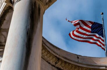 Perspectiva en contrapicado de la bandera de EEUU ondeando con firmeza entre columnas clásicas de piedra en Salt Lake City (Utah, EEUU), perfilada contra un cielo azul sin nubes. Dependencias