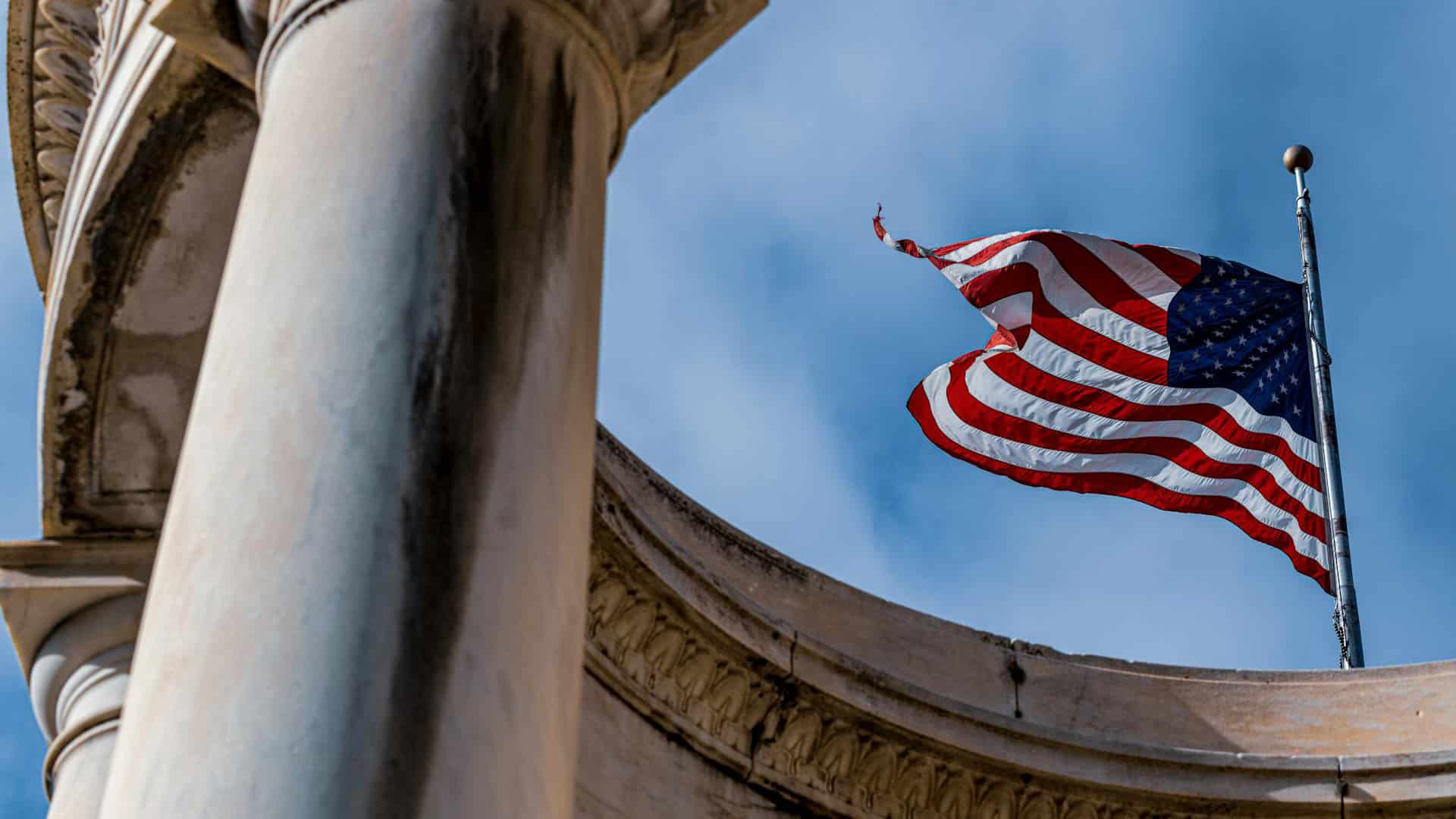 Perspectiva en contrapicado de la bandera de EEUU ondeando con firmeza entre columnas clásicas de piedra en Salt Lake City (Utah, EEUU), perfilada contra un cielo azul sin nubes. Dependencias