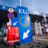 A wide-angle view of the Calafat crossing between Bulgaria and Romania: in the foreground, a blue sign bearing the EU emblem and a white arrow; beyond it, control booths, red-and-white retractable barriers, speed-limit signs, and an attendant in a blue jacket walking away under a clear sky. Schengen