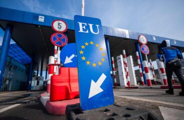 A wide-angle view of the Calafat crossing between Bulgaria and Romania: in the foreground, a blue sign bearing the EU emblem and a white arrow; beyond it, control booths, red-and-white retractable barriers, speed-limit signs, and an attendant in a blue jacket walking away under a clear sky. Schengen
