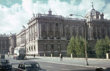 The Royal Palace of Madrid (Spain) in the 1960s. A large, historic building with classical architecture, featuring ornate details and a dome on the right side. The building is situated along a street with several old cars and a double-decker bus. Trees are visible in front of the building, and the sky is partly cloudy.