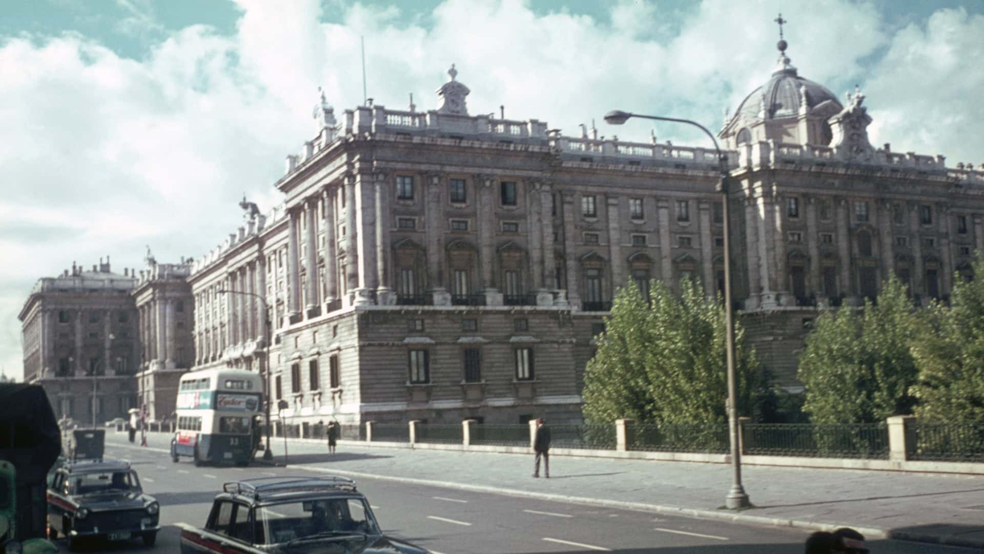 The Royal Palace of Madrid (Spain) in the 1960s. A large, historic building with classical architecture, featuring ornate details and a dome on the right side. The building is situated along a street with several old cars and a double-decker bus. Trees are visible in front of the building, and the sky is partly cloudy.