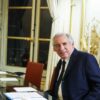 François Bayrou, primer ministro de Francia, sentado en una mesa de reuniones con documentos, portátil y vasos de agua, en una sala elegante con cortinas blancas y detalles dorados con banderas de Francia y la Unión Europea al fondo. Imagen recortada del encuentro con Magnus Brunner, comisario europeo de Asuntos Internos y Migración, en París. (20/01/2025).