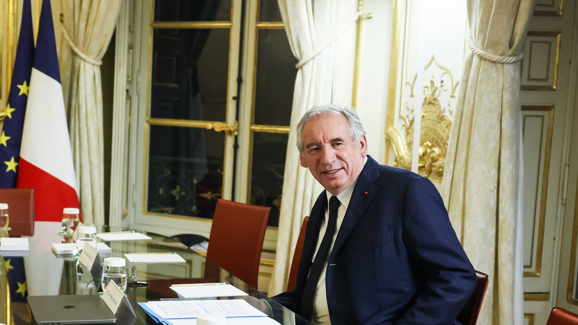 François Bayrou, primer ministro de Francia, sentado en una mesa de reuniones con documentos, portátil y vasos de agua, en una sala elegante con cortinas blancas y detalles dorados con banderas de Francia y la Unión Europea al fondo. Imagen recortada del encuentro con Magnus Brunner, comisario europeo de Asuntos Internos y Migración, en París. (20/01/2025).