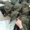 Jóvenes (entre ellos inmigrantes) aspirantes a soldados profesionales del Ejército de Tierra en el Centro de Formación de Tropa nº 2 (2019) con uniformes de camuflaje revisando un mapa desplegado en el suelo; uno de ellos señala una zona del mapa mientras portan mochilas y rifles.
