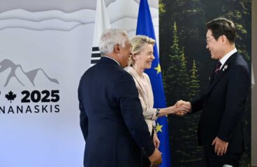 Handshake between Lee-Jae Myung, President of South Korea (on the right), and Ursula Von der Leyen, in the presence of Antonio Costa (on the left). They are standing in front of the flags of South Korea and the European Union, with a background sign reading ‘G7 2025 KANANASKIS’ featuring mountains and a maple leaf.